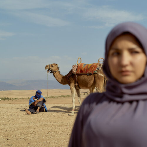 A young woman in a hijab stands in focus with a blurred background featuring a man and a camel in a desert landscape, Morocco