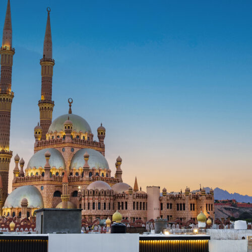A stunning mosque stands with its tall minarets reaching towards the evening sky, surrounded by tranquil desert scenery and mountains in the background. The scene captures a sense of peace.